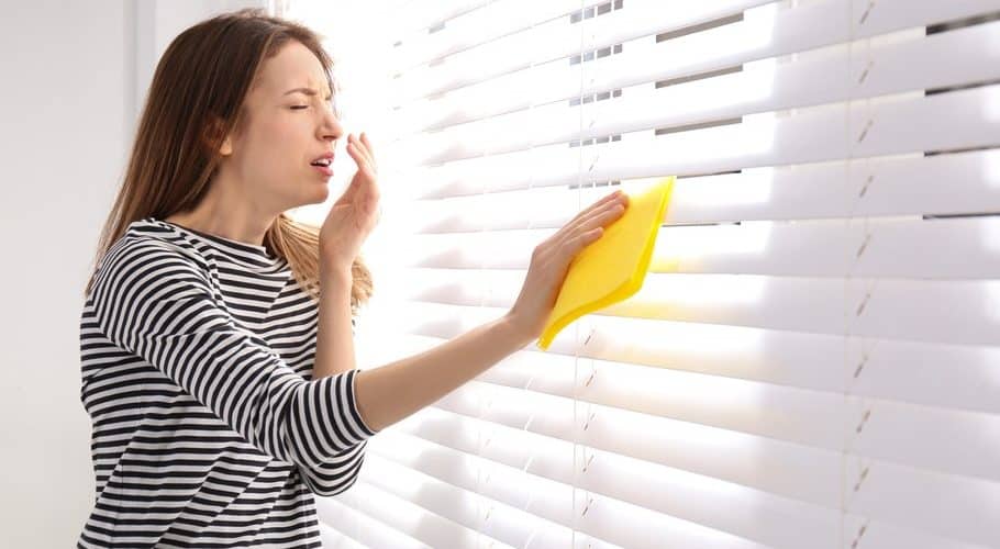 woman sneezing while dusting window blinds with dust and allergens in the air